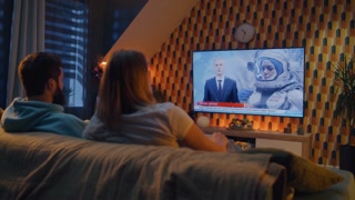 Couple sitting on sofa with snacks in living room, watching news on TV channel about astronauts, space mission, exploring Earth orbit. Wife and husband resting and chilling at home on weekend at night