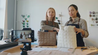 Medium shot of leather workshop owner and employee filming story or reel on smartphone for social media about new handbag, and demonstrating its features for subscribers