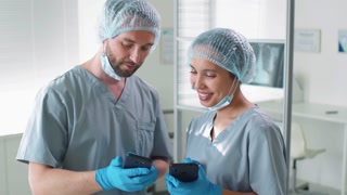 Male and female hospital workers in protective uniforms smiling and chatting while watching social media on their smartphones during break